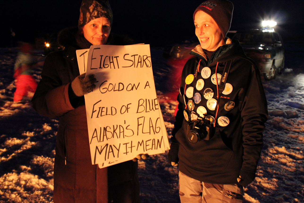 At the closing ceremonies of the 2014 Homer Winter Carnival on Saturday, Cathy Essex holds cue cards with the lyrics of Alaska’s Flag Song. Nichole Essex, right, displays her collection of  HWC buttons.-Photo by McKibben Jackinsky, Homer News