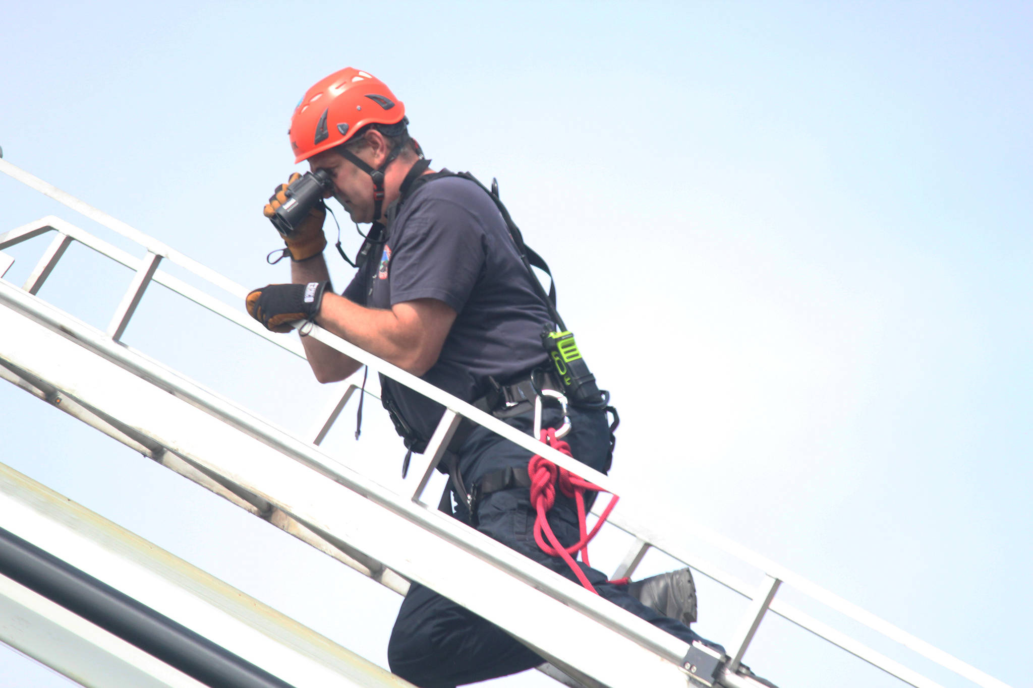 Joe Sallee, Deputy Chief at Kachemak Emergency Services, uses binoculars at the end of a ladder extended from the KES ladder truck to search the slope near Baycrest Overlook on Monday, July 24, 2017 in Homer, Alaska. First responders found a pickup truck that had fallen over the edge after hitting a guard rail, and searched for a possible victim until getting a report that the driver had crashed several days earlier and had walked away uninjured.  Photo by Megan Pacer/Homer News