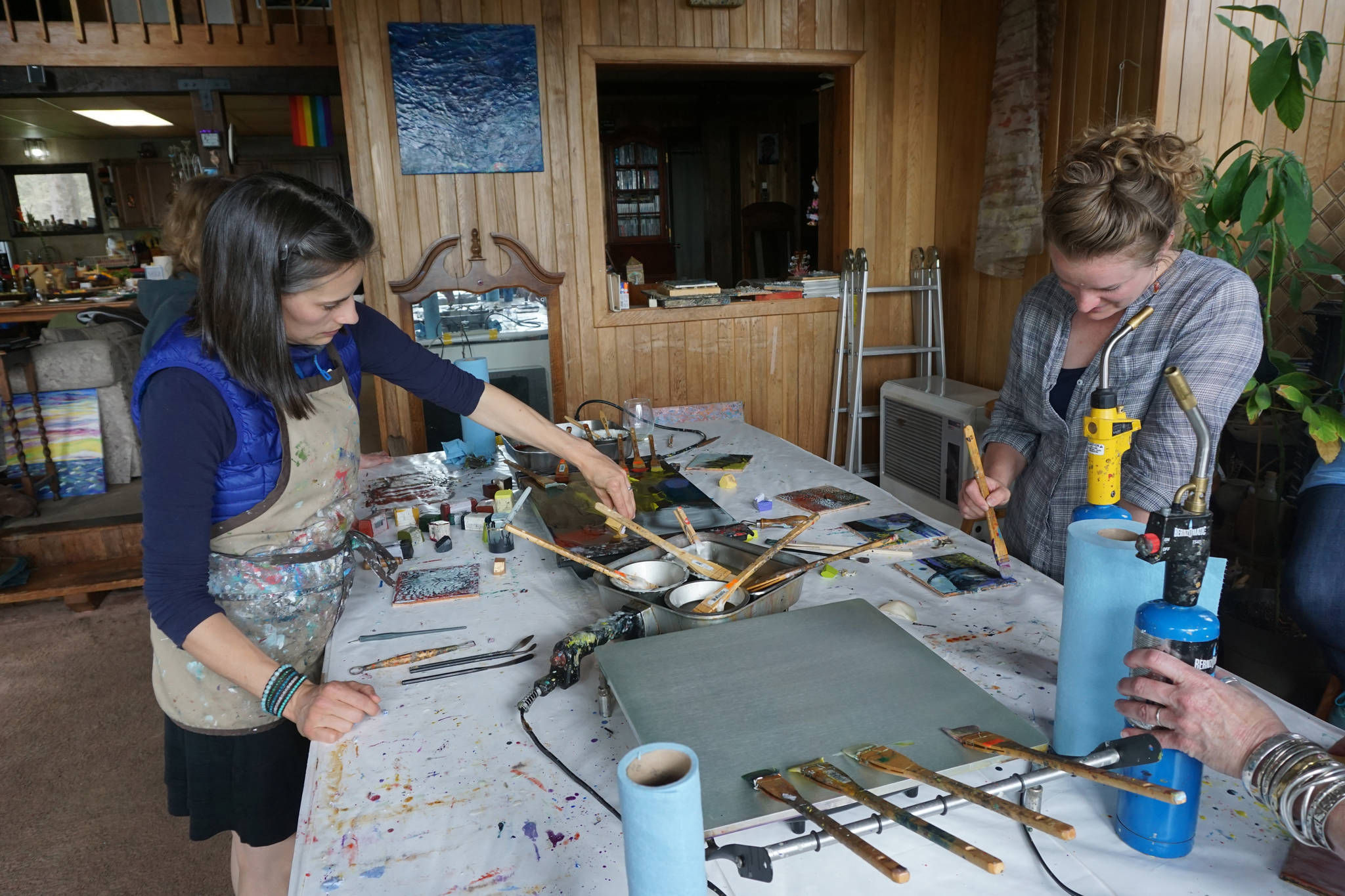 Carla Klinker-Cope., left, and Becca Bottebaum paint with encaustics in Ann-Margret Wimmerstedt’s “Wax. Wine & Wimmerstedt”class held July 30, 2017 at Wimmerstedt’s home. Wimmerstedt, far right, reaches for a propane torch. The torch is used to manipulate the wax by melting it and also to fuse the finished painting. “Encaustic” means “to burn.” (Photo by Michael Armstrong/Homer News)
