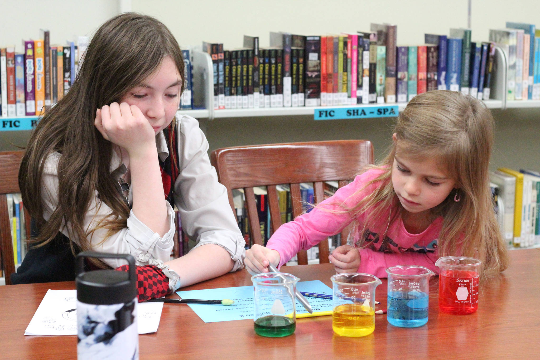 Jade Stewart (left) watches her mentee from Paul Banks Elementary School, Jillian Koran, perform an experiment at one of five stations Tuesday, Oct. 24, 2017 at Homer Middle School in Homer, Alaska. First grade students were paired up with eighth graders for a lesson on the science of sound through a mentorship program. (Photo by Megan Pacer/Homer News)
