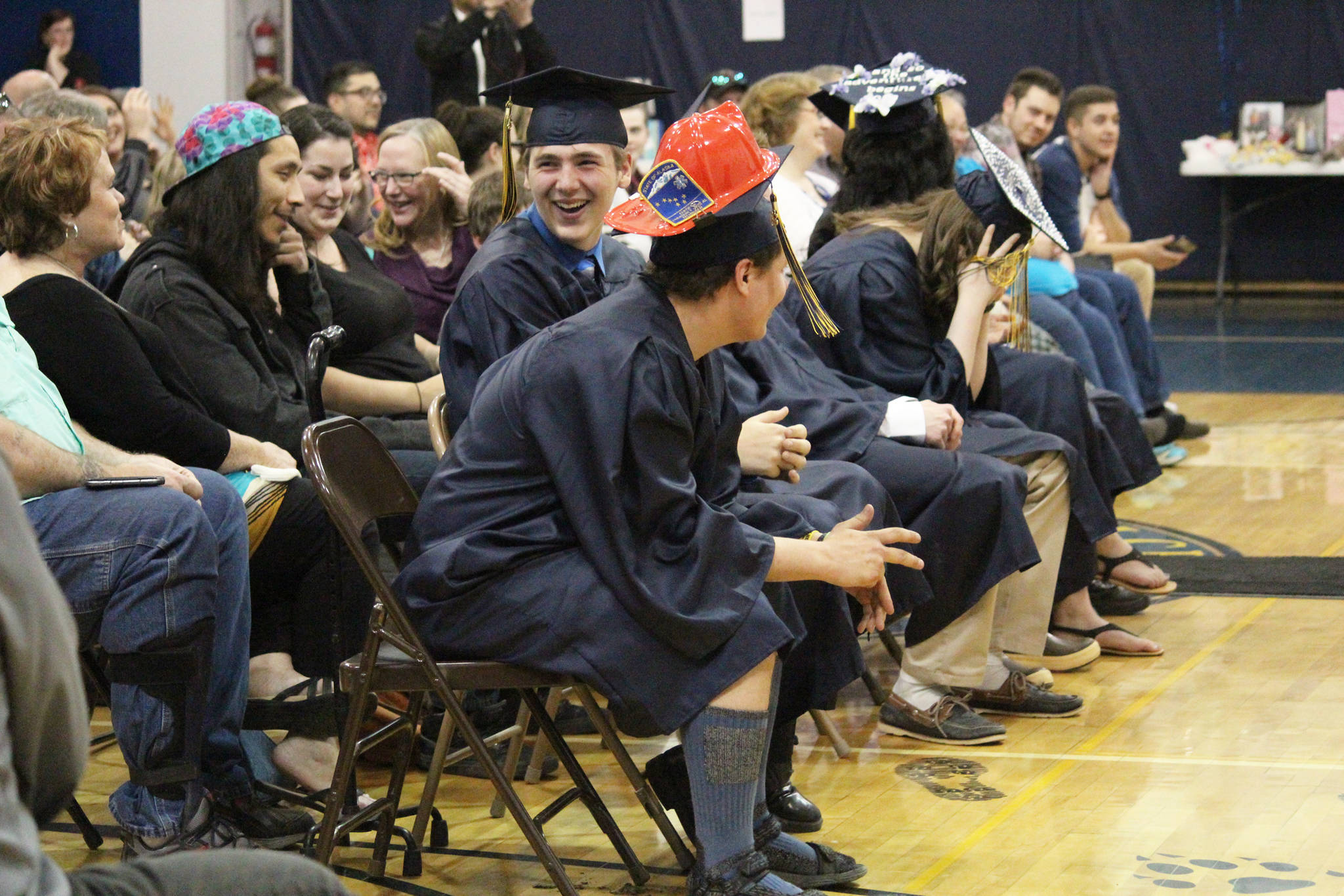 Members of the Ninilchik School graduating class of 2018 laugh and react as one of the speakers at their Monday, May 21, 2018 ceremony walks back to his seat at the school in Ninilchik, Alaska. The speaker accidentally took a page of salutatorian Chelsea Oberle-Lozano’s speech from the podium after finishing his own. (Photo by Megan Pacer/Homer News)