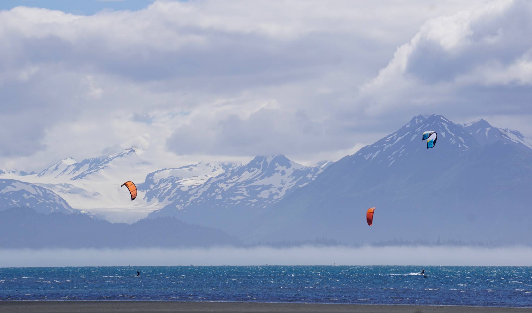 Kite boarders play in Kachemak Bay while a fog bank covers the south shore last Saturday afternoon, June 30, 2018, in Homer, Alaska. When the afternoon bay breeze kicks in, Homer’s kite boarding community can be seen off the Mariner Park beach. (Photo by Michael Armstrong/Homer News)