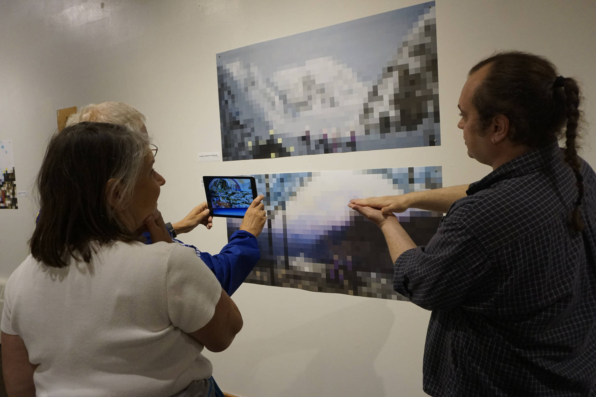 James Dolma views an image in an iPad from the “Dirigibles of Denali” show at the Pratt Museum at the July 6, 2018 First Friday opening in Homer, Alaska. The images appear when viewed through the HP Reveal augmented reality app. Artist Nathan Shafer, far right, explains the process to Brenda Dolma, center. (Photo by Michael Armstrong/Homer News)