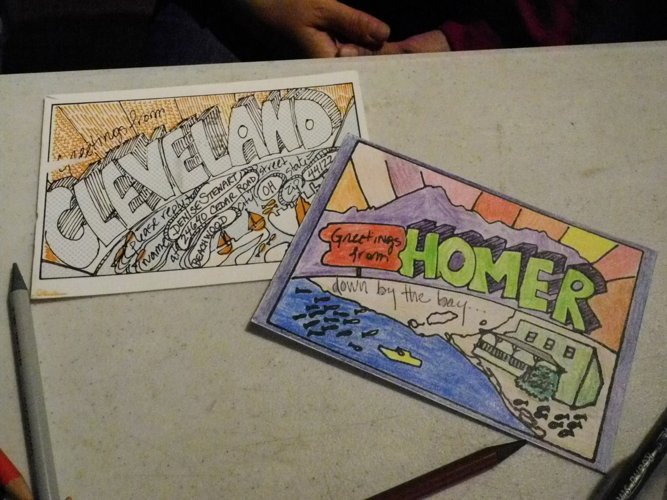 A “Greetings from Homer” postcard colored by Jenny Stroyeck of Homer is next to a Cleveland card. Emery encouraged Homer residents to visit Bunnell Street Arts Center and color Homer postcards to exchange with Cleveland area residents.-Photo by Michael Armstrong, Homer News