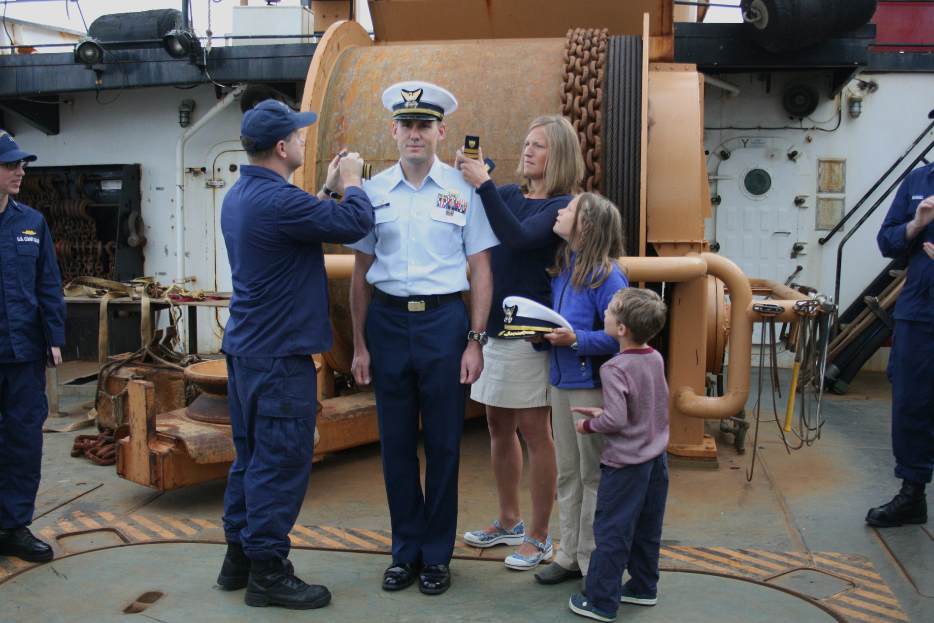 Chief Petty Officer Travis Clark assists the family of Commander Brian C. Krautler in pinning on Krautler’s new shoulder boards bearing the Commander (O-5) insignia. Krautler, the commanding officer of the Hickory, received the promotion effective July 1. Pictured are Clark, Krautler, Krautler’s wife, Andrea, and their children, Kate, 9,  and James, 5.  The promotion was celebrated in a small ceremony held aboard the Hickory at its berth on Pioneer Pier.