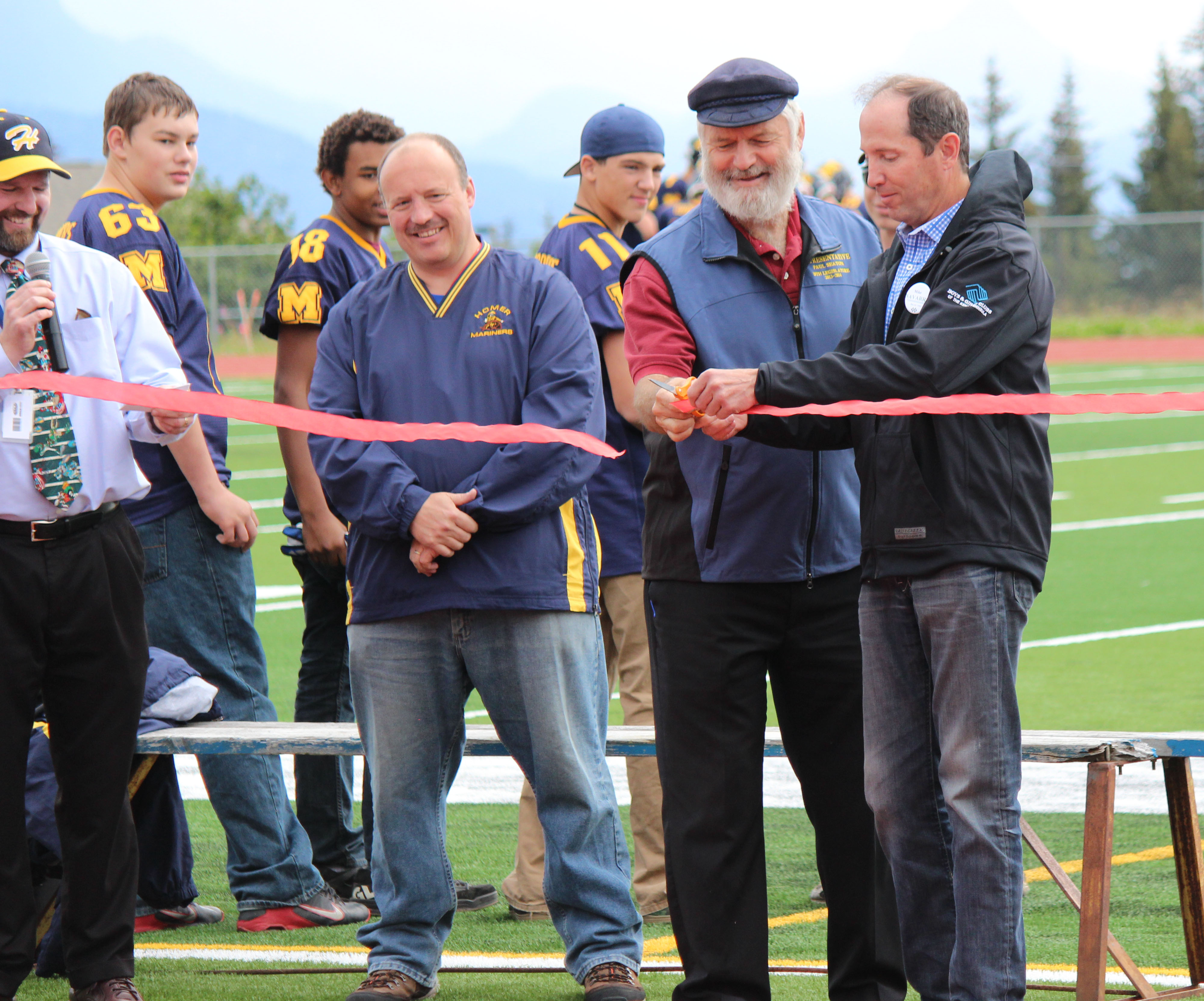 Rep. Paul Seaton, second from right, and Kenai Peninsula Borough Mayor Mike Navarre, right, cut the ribbon at the dedication ceremony of Homer High School’s new turf field on Saturday. Looking on are HHS Principal Doug Waclawski, left, and Kenai Peninsula Borough School District Assistant Superintendent Sean Dusek, with members of the Mariners football team in the background. Exhibitions of different sports that will use the field took place during halftime of the Mariners’ Satuarday game against Eielson.