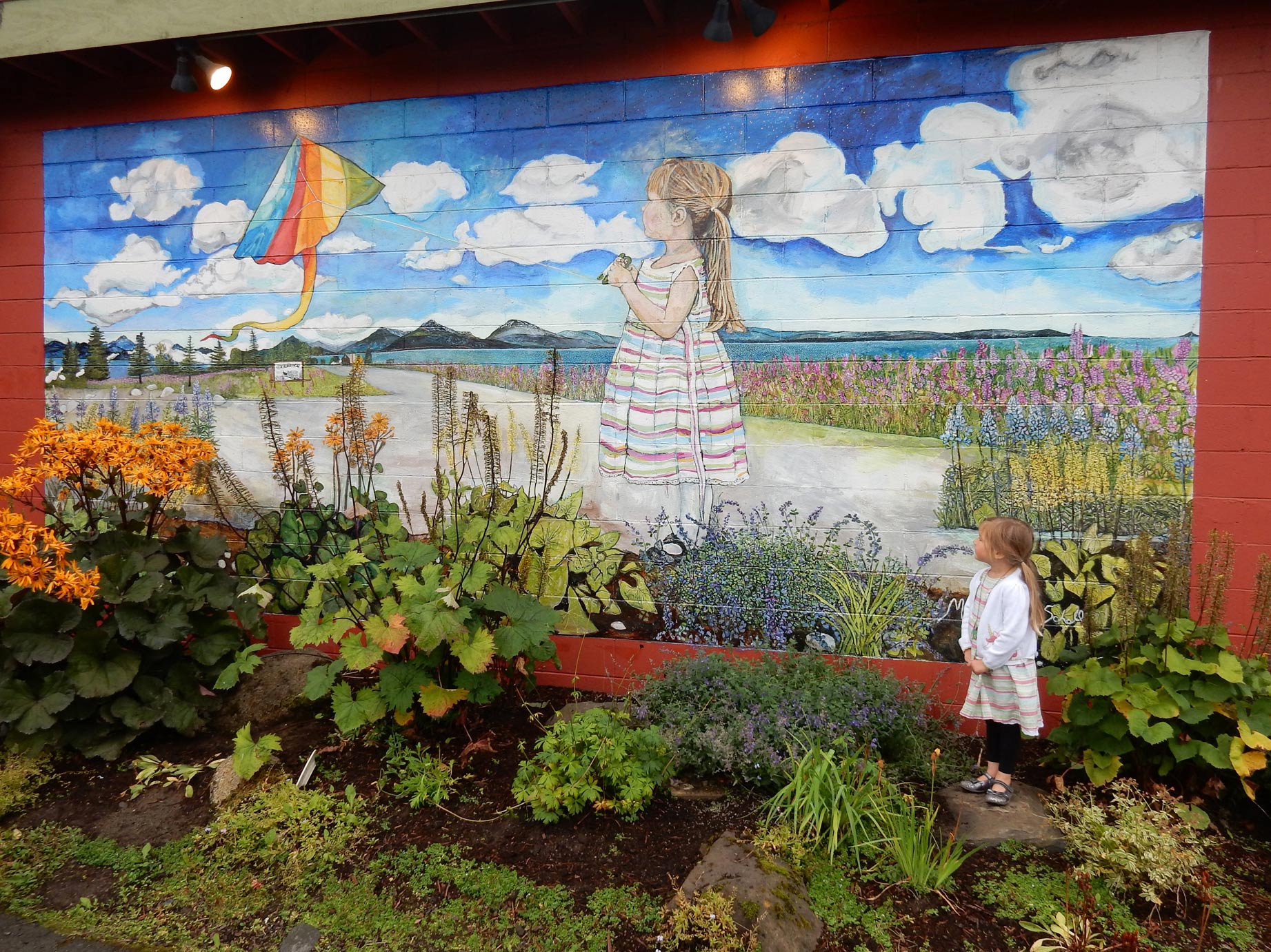 Morgan Harness, who just celebrated her 4th birthday, poses beside the Marjorie Scholl mural, in which she is pictured flying a kite. The mural, which was unveiled Friday on the side of Fat Olives is one of the last works to be installed under a $150,000 ArtPlace America grant that funded public art and artists’ residencies in Old Town.-Photo by Mercedes Harness