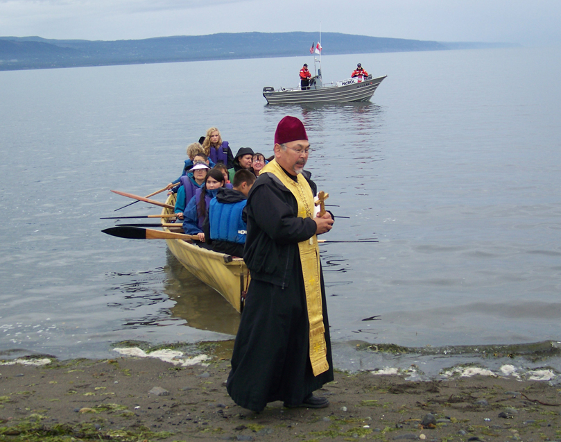 Father Thomas Andrew of the Holy Assumption of the Virgin Mary Russian Orthodox Church in Kenai, offers a blessing on the arriving kayaks, as members of the Coast Guard Auxiliary’s Homer flotilla keep watch.-Photo by McKibben Jackinsky, Homer News