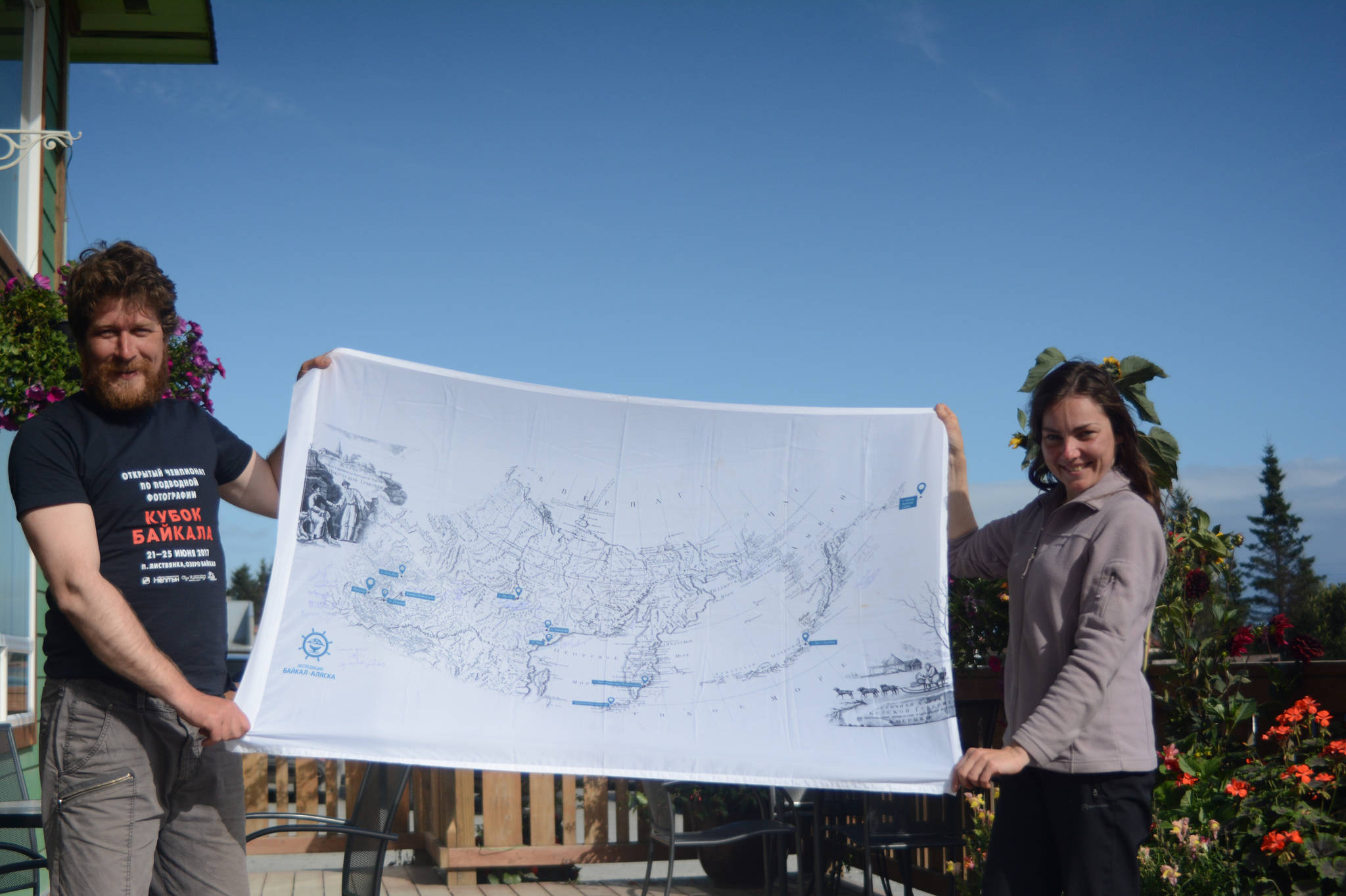 Anatoly Kazakevich, left, and Anna Vazhenina , right, hold up a flag showing the route of the voyage of their inflatable catamaran, Iskatel, last Thursday, Aug. 30, 2018, at K-Bay Caffe in Homer, Alaska. They finished sailing from Lake Baikal, Russia, to Homer, on Aug. 26. The flag is from an illustration from the 1803-06 voyage of Capt. Ivan Kruzenshtern. (Photo by Michael Armstrong/Homer News)