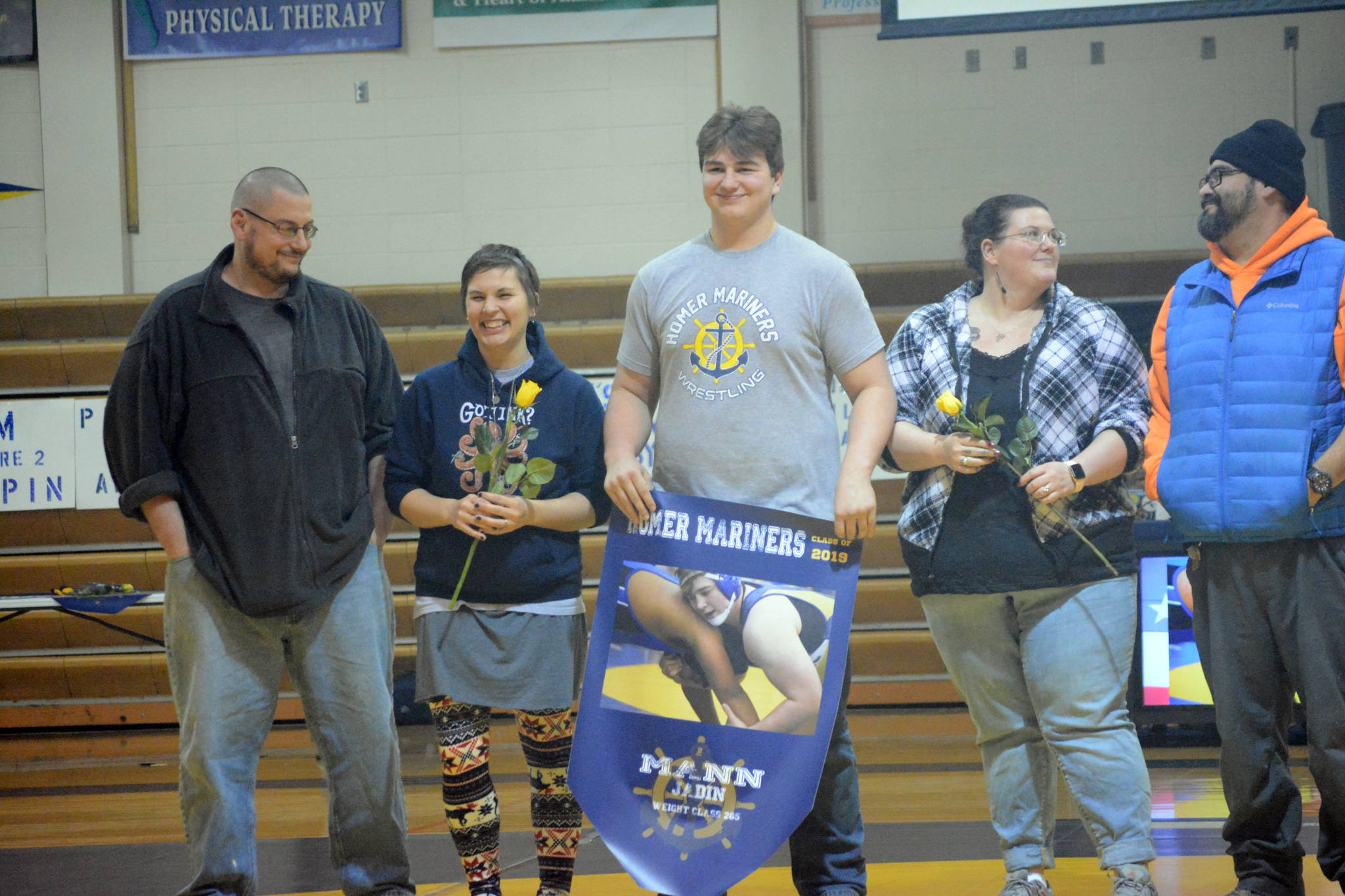 Jadin Mann holds a banner recognizing him at Senior Night, the Homer-Kenai dual wrestling meet held Nov. 21, 2018, in the Homer High School Alice Witte Gym, Homer, Alaska. Supporting him are his parents, Danny and Abi Rios, right??, and Brian and Heather Mann, left??, (Photo by Michael Armstrong/Homer News)