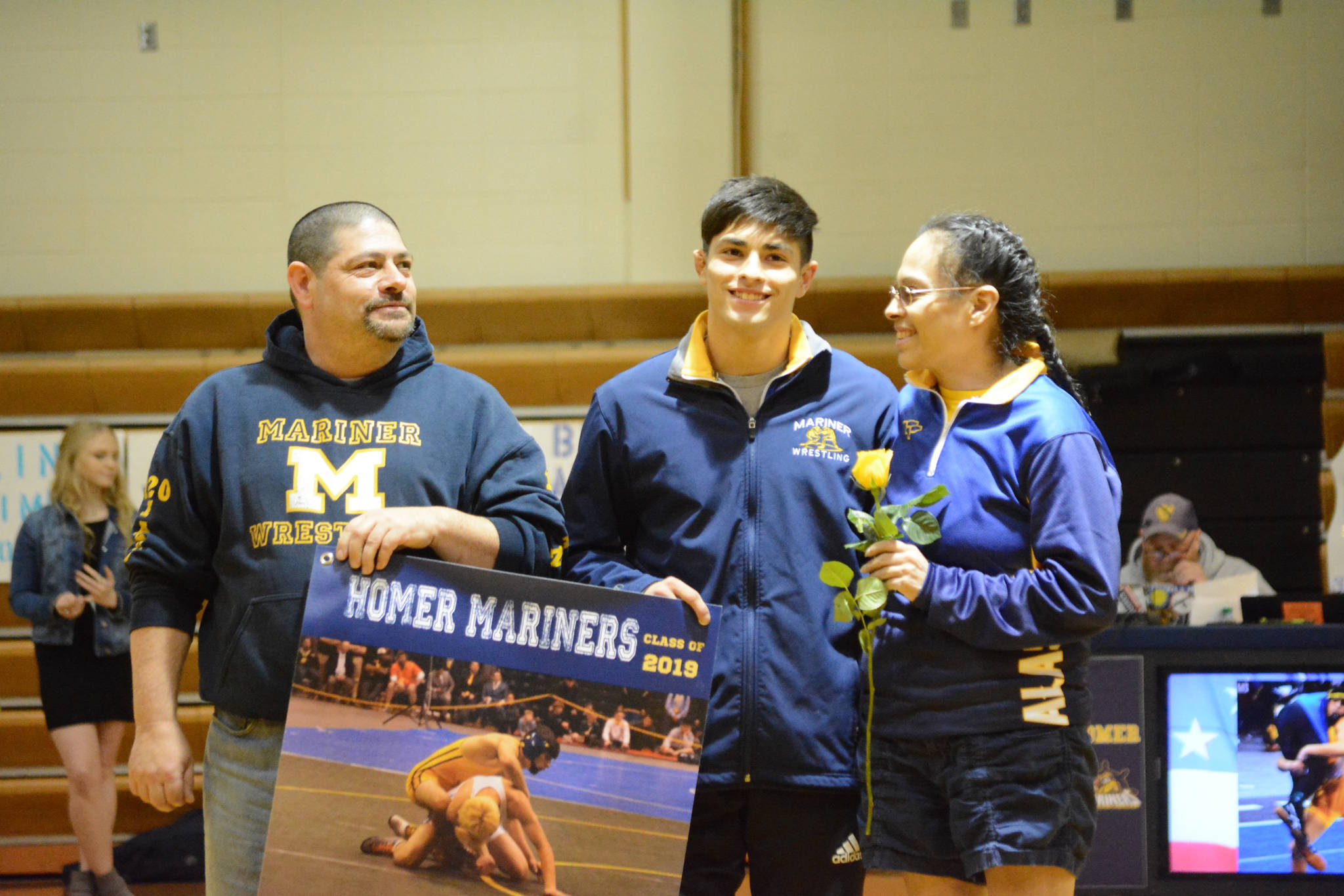 Luciano Fasulo holds a banner recognizing him at Senior Night, the Homer-Kenai dual wrestling meet held Nov. 21, 2018, in the Homer High School Alice Witte Gym, Homer, Alaska. Supporting hi are Greg and Janie Martin. (Photo by Michael Armstrong/Homer News)