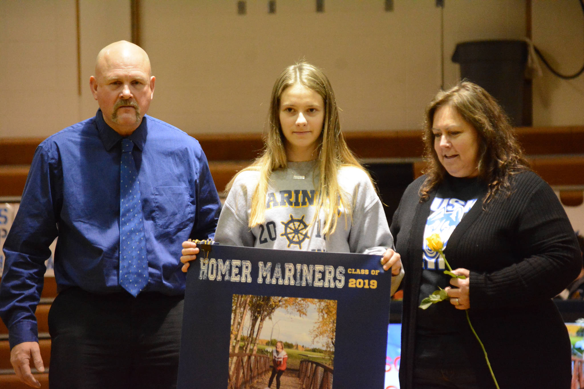 Allison Wells holds a banner recognizing her at Senior Night, the Homer-Kenai dual wrestling meet held Nov. 21, 2018, in the Homer High School Alice Witte Gym, Homer, Alaska. Supporting her are her parents, Bubba and Mike Wells.(Photo by Michael Armstrong/Homer News)