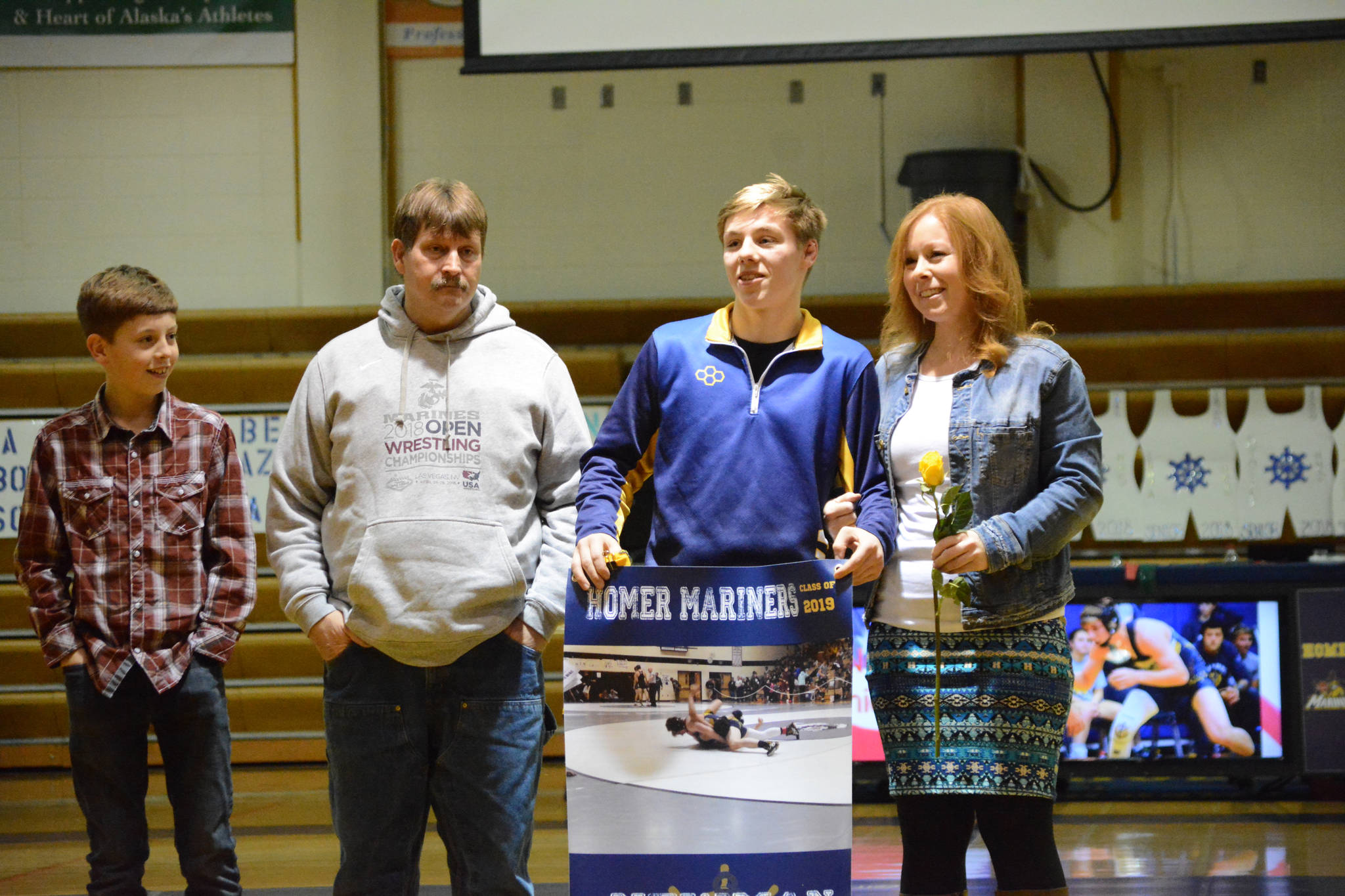 Wayne Newman holds a banner recognizing him at Senior Night, the Homer-Kenai dual wrestling meet held Nov. 21, 2018, in the Homer High School Alice Witte Gym, Homer, Alaska. Supporting him are his parents, Sam and Auriell Newman, and brother, ??? Newman. (Photo by Michael Armstrong/Homer News)