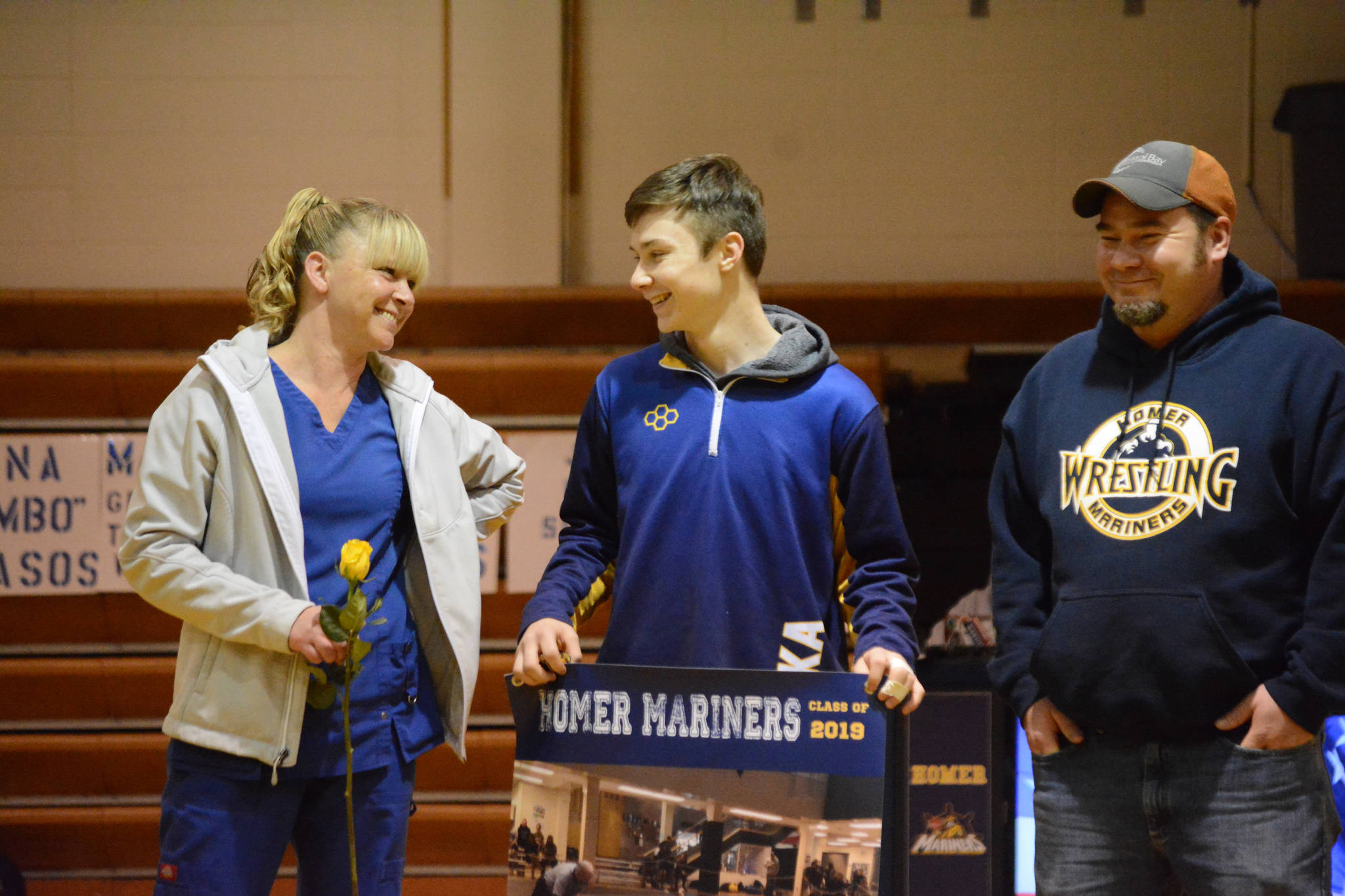 Ian Stovall holds a banner recognizing him at Senior Night, the Homer-Kenai dual wrestling meet held Nov. 21, 2018, in the Homer High School Alice Witte Gym, Homer, Alaska. Supporting him are his parents, Katie Watson and Will Stovall. (Photo by Michael Armstrong/Homer News)