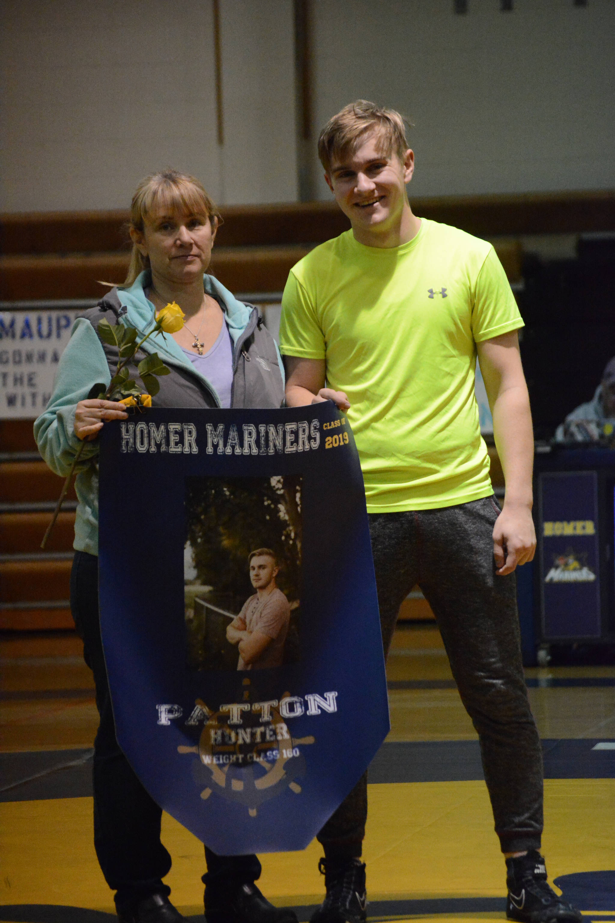 Hunter Patton holds a banner recognizing him at Senior Night, the Homer-Kenai dual wrestling meet held Nov. 21, 2018, in the Homer High School Alice Witte Gym, Homer, Alaska. Supporting him is his mother, Lyn Patton, and father, Tom Patton, not shown. (Photo by Michael Armstrong/Homer News)