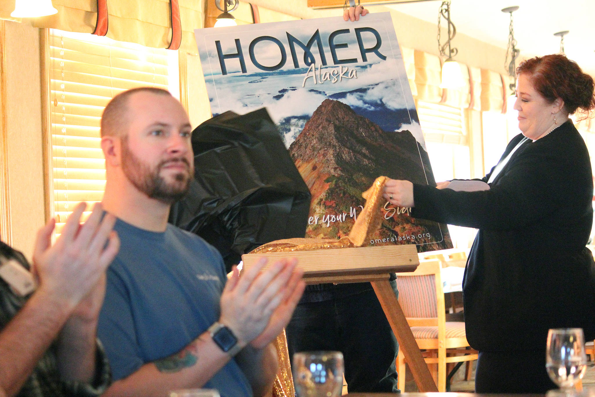 Homer Chamber of Commerce Executive Director Debbie Speakman unveils the winning photo that will grace the cover of the chamber’s next visitor guide at a Tuesday, Jan. 15, 2019 annual chamber meeting at the Best Western in Homer, Alaska. Russel Campbell of Wandering Nomad Photography took the photo. (Photo by Megan Pacer/Homer News)
