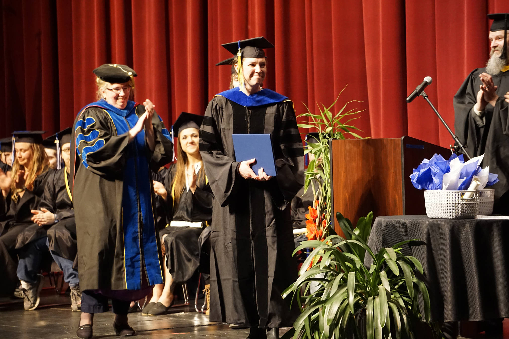 Emilie Springer smiles after receiving her doctor in anthropology degree and hood at the Kachemak Bay Campus commencement last Wednesday, May 8, 2019, at the Mariner Theatre in Homer, Alaska. Applauding at left is KBC interim campus director Paula Martin. A Homer resident from Ninilchik, Springer received her doctorate from the University of Alaska Fairbanks but chose to receive her PhD. in her home town — the first doctorate to do so at KBC commencement. (Photo by Michael Armstrong/Homer News.)