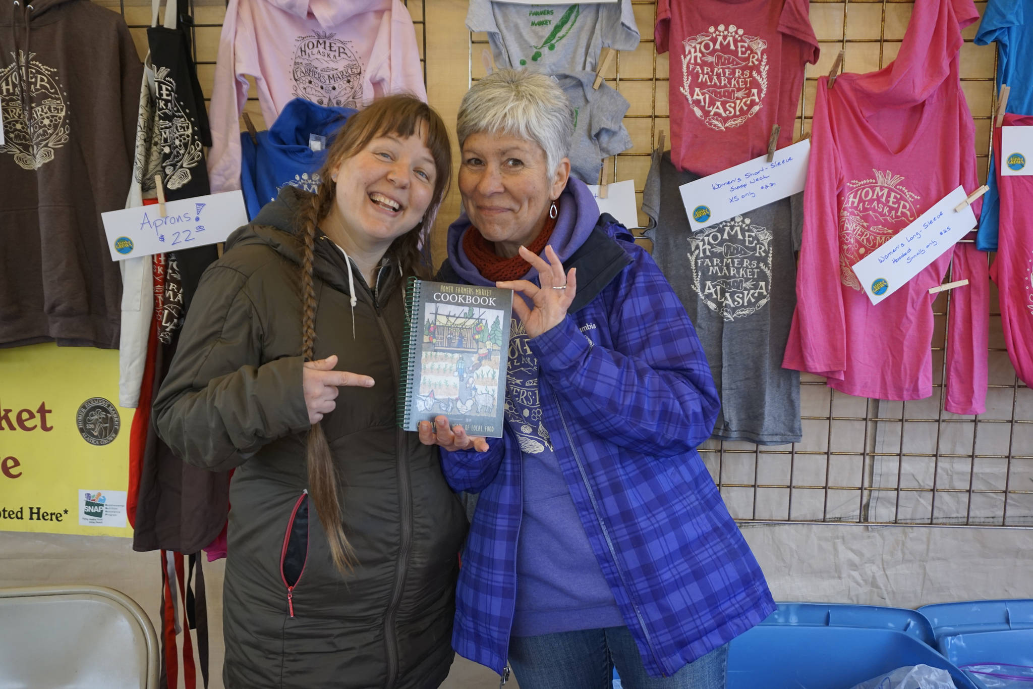 Homer Farmers Market Director Robbi Mixon, left, and Margarida Kondak, right, display of copy of the Homer Farmers Market Cookbook at the opening of the market on May 25, 2019, in Homer, Alaska. (Photo by Michael Armstrong/Homer News)