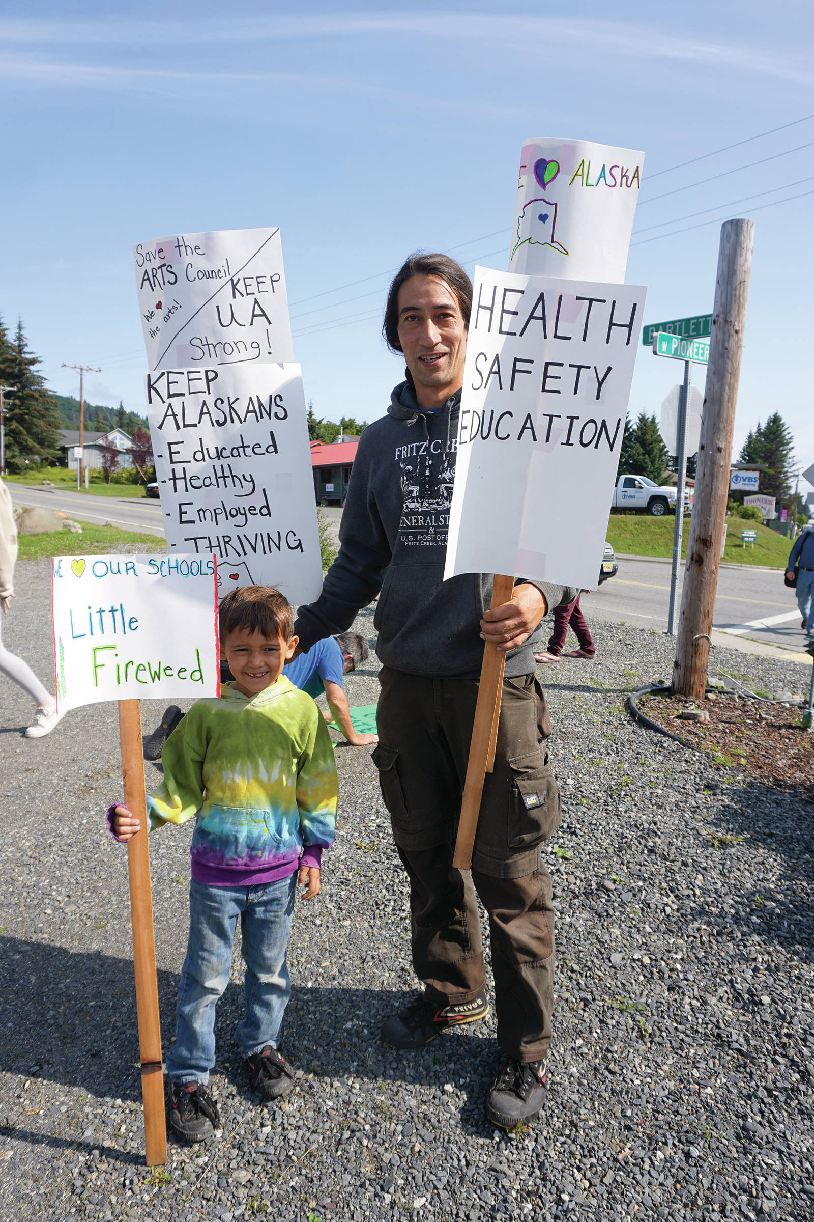 Nako Cook, left and Jared Cook, right, hold signs at a rally Sunday, July 28, 2019, against Gov. Mike Dunleavy’s budget cuts at the Legislative Information Office, Homer, Alaska. (Photo by Michael Armstrong/Homer News).