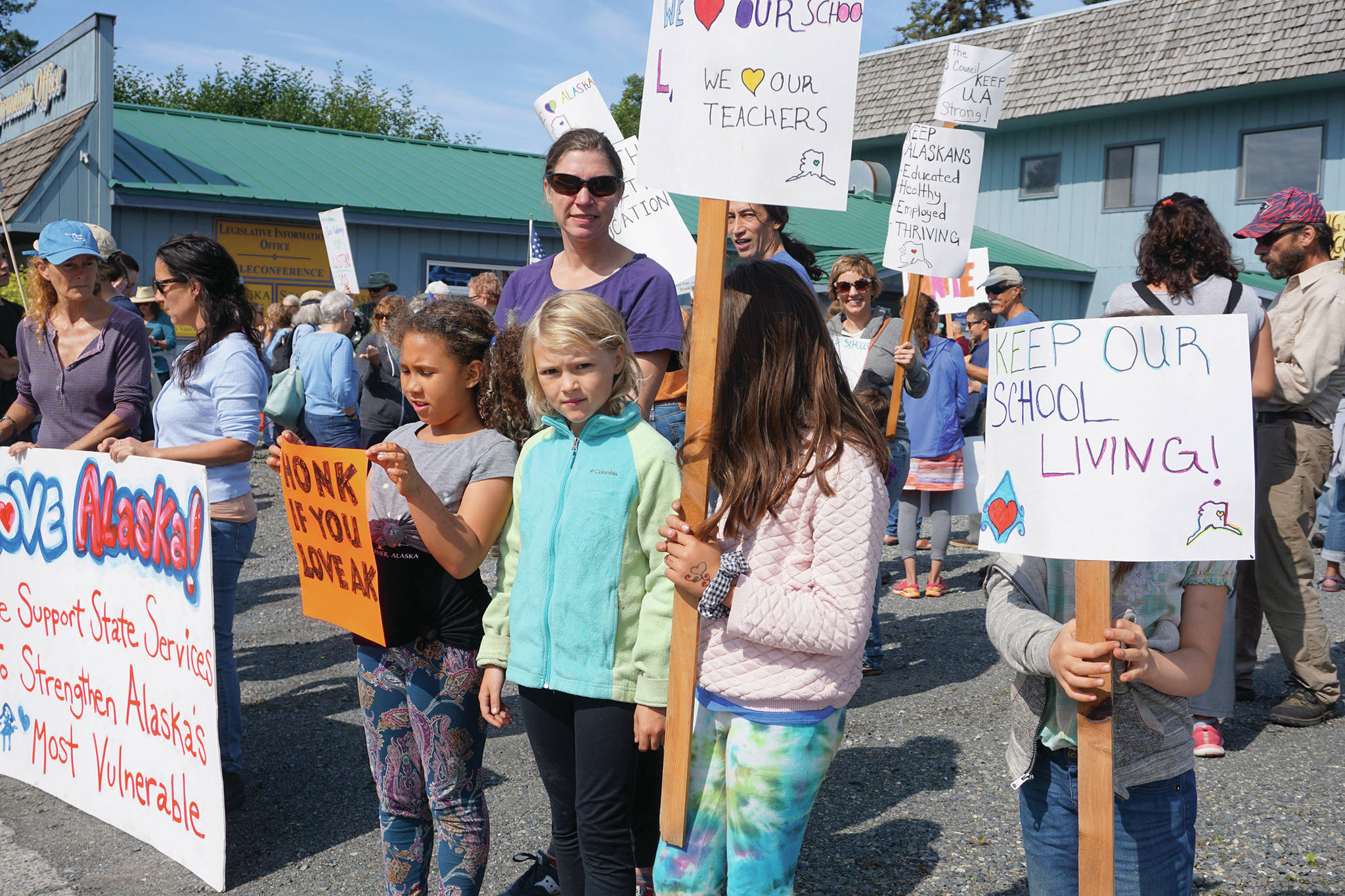 Demonstrators of all ages hold up signs at a rally Sunday, July 28, 2019, against Gov. Mike Dunleavy’s budget cuts at the Legislative Information Office, Homer, Alaska. (Photo by Michael Armstrong/Homer News).