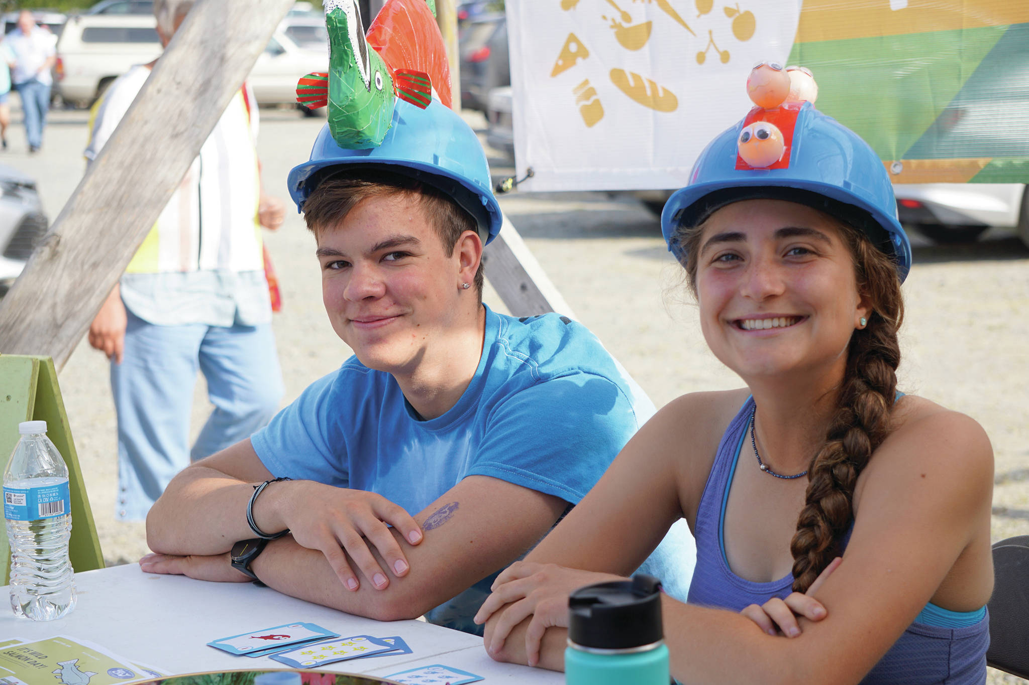 Eli Arbogast, left, and Rosie Skovron, right, staff a table on Saturday, Aug. 10, 2019, for Alaska Wild Salmon Day at the Homer Farmers Market in Homer, Alaska. Arbogast wears a salmon hat and Skovron a hat with “a row of roe,” she said. They are summer interns at Cook Inletkeeper. (Photo by Michael Armstrong/Homer News)