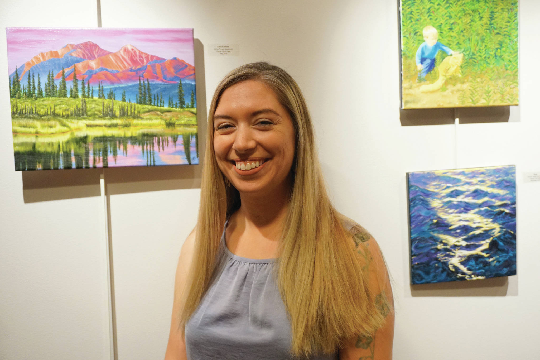 Maria Bernier poses by some of her paintings at her show at Kachemak Bay Campus that opened on First Friday, Oct. 4, 2019, in Homer, Alaska. (Photo by Michael Armstrong/Homer News)