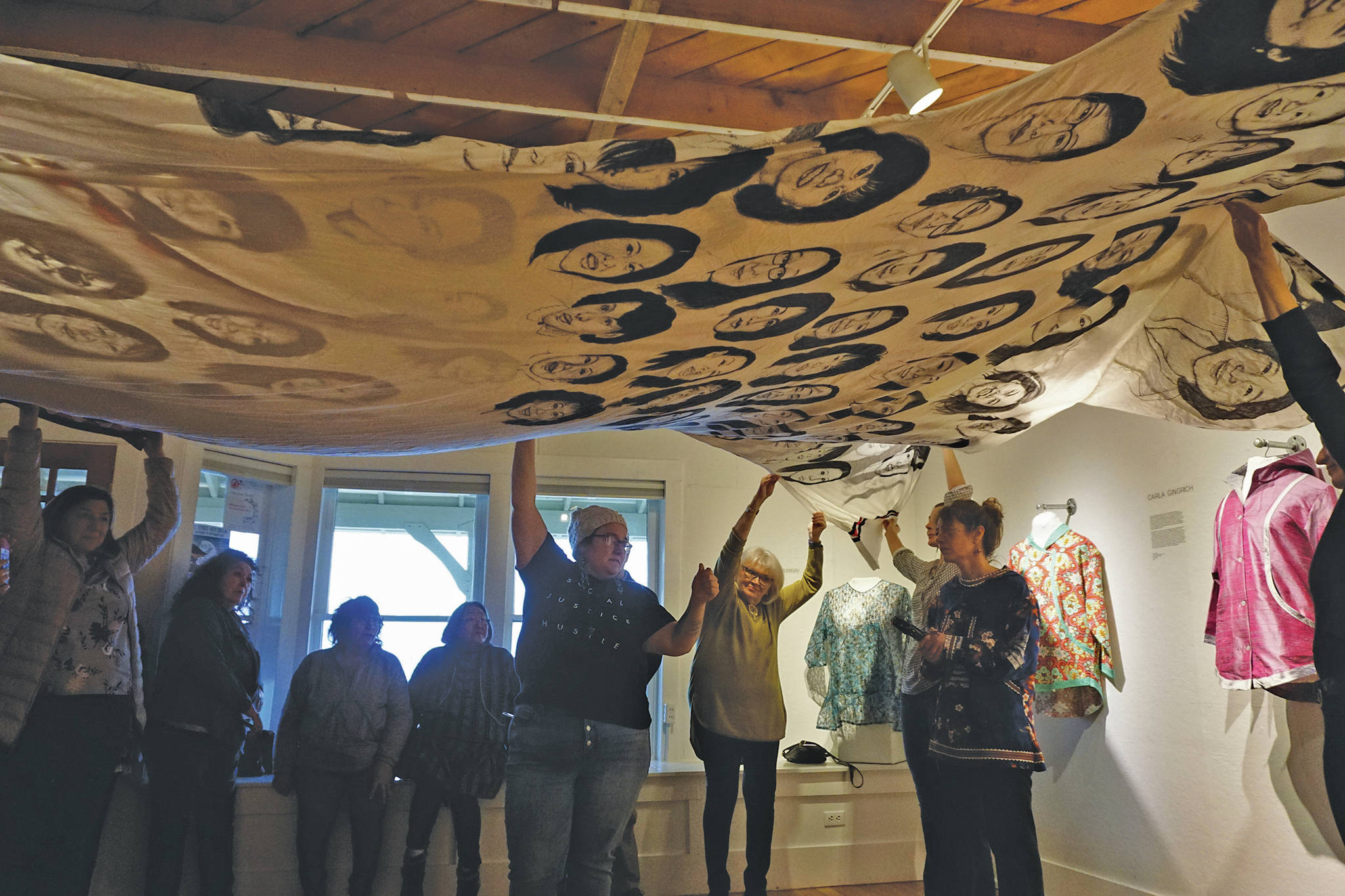 First Friday visitors hold up Amber Webb’s kuspuk at the opening of the “Kuspuk/Qaspeq/Atikluk” show on Oct. 5, 2019, at Bunnell Street Arts Center in Homer, Alaska. The kuspuk shows faces of missing or murdered indigenous women. (Photo by Michael Armstrong/Homer News)