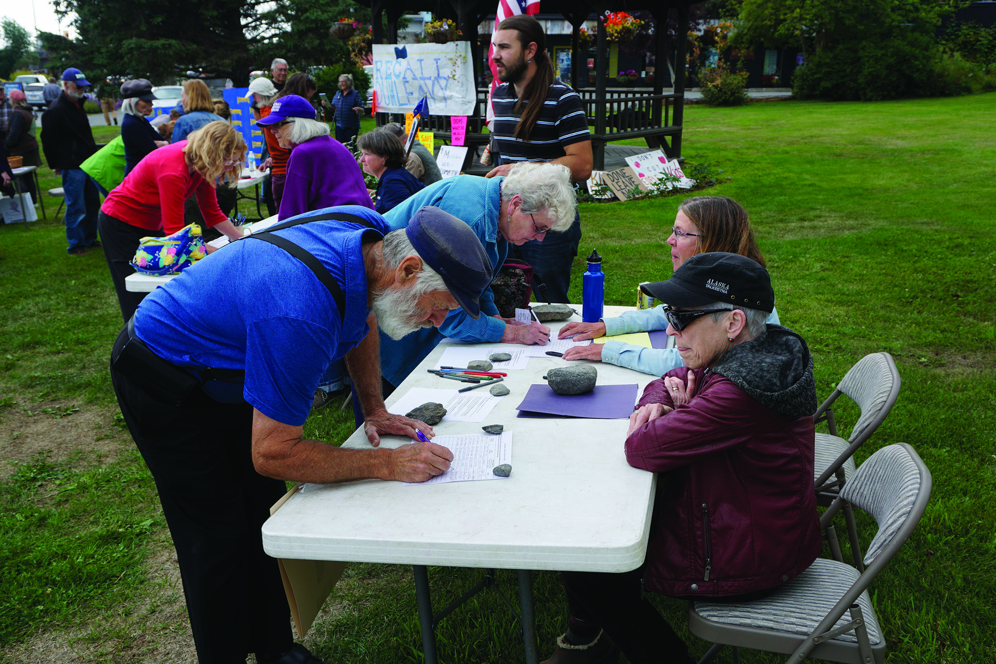 Recall Dunleavy organizers Ann Keffer, right, in hat, and Pat Cue, behind Keffer, take signatures at a Recall Dunleavy rally held on Aug. 1, 2019, at WKFL Park in Homer, Alaska. At left, former Homer Rep. Paul Seaton, NP-Homer, signs a form. Seaton lost to Rep. Sarah Vance, R-Homer, in the general election. (Photo by Michael Armstrong)