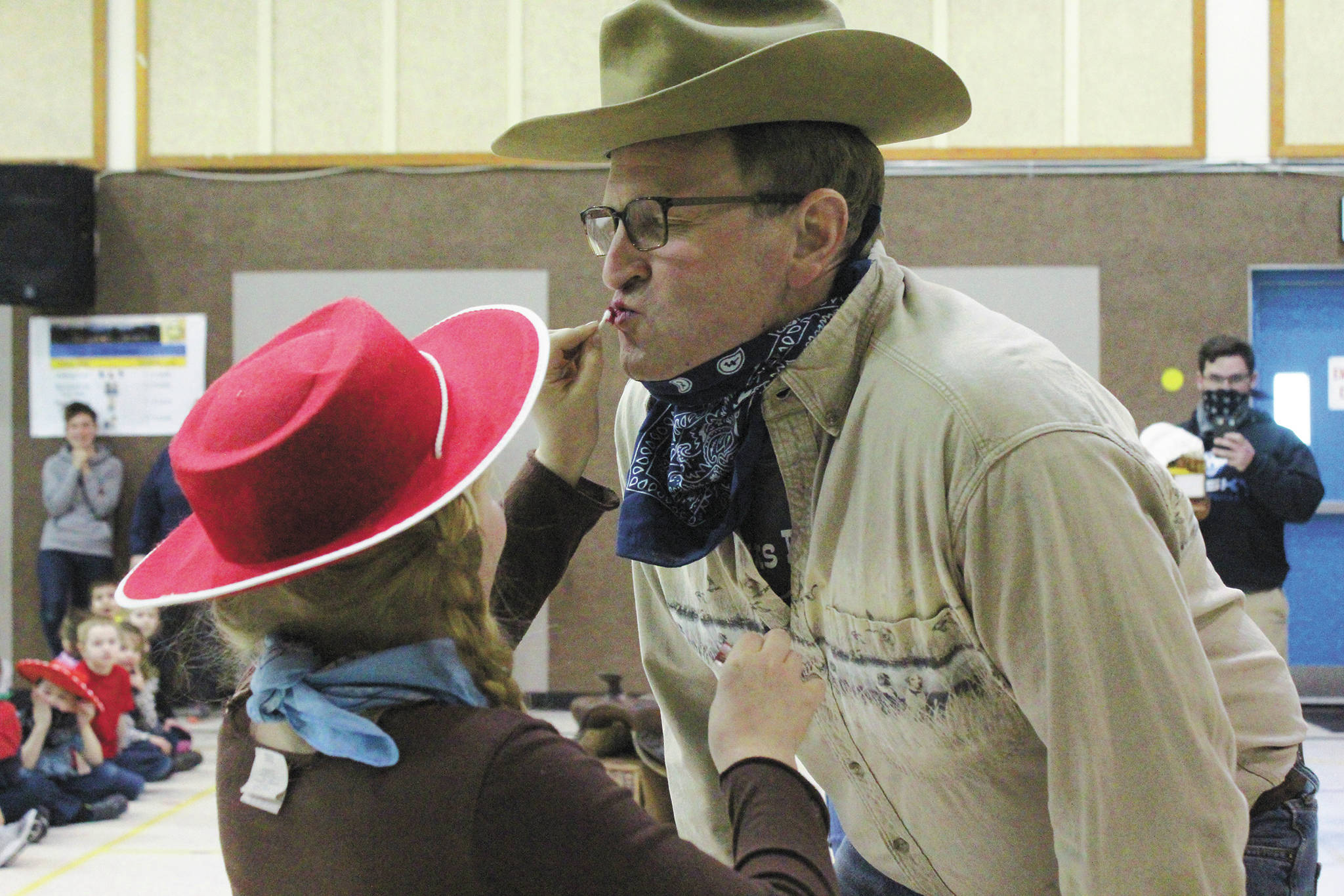 Ingrid Pederson, Principal Eric Pederson’s daughter, helps him put on lipstick before he kisses a pig at a wild west themed assembly to celebrate the end of Paul Banks Elementary School’s annual read-a-thon Thursday, March 5, 2020 at the school in Homer, Alaska. (Photo by Megan Pacer/Homer News)