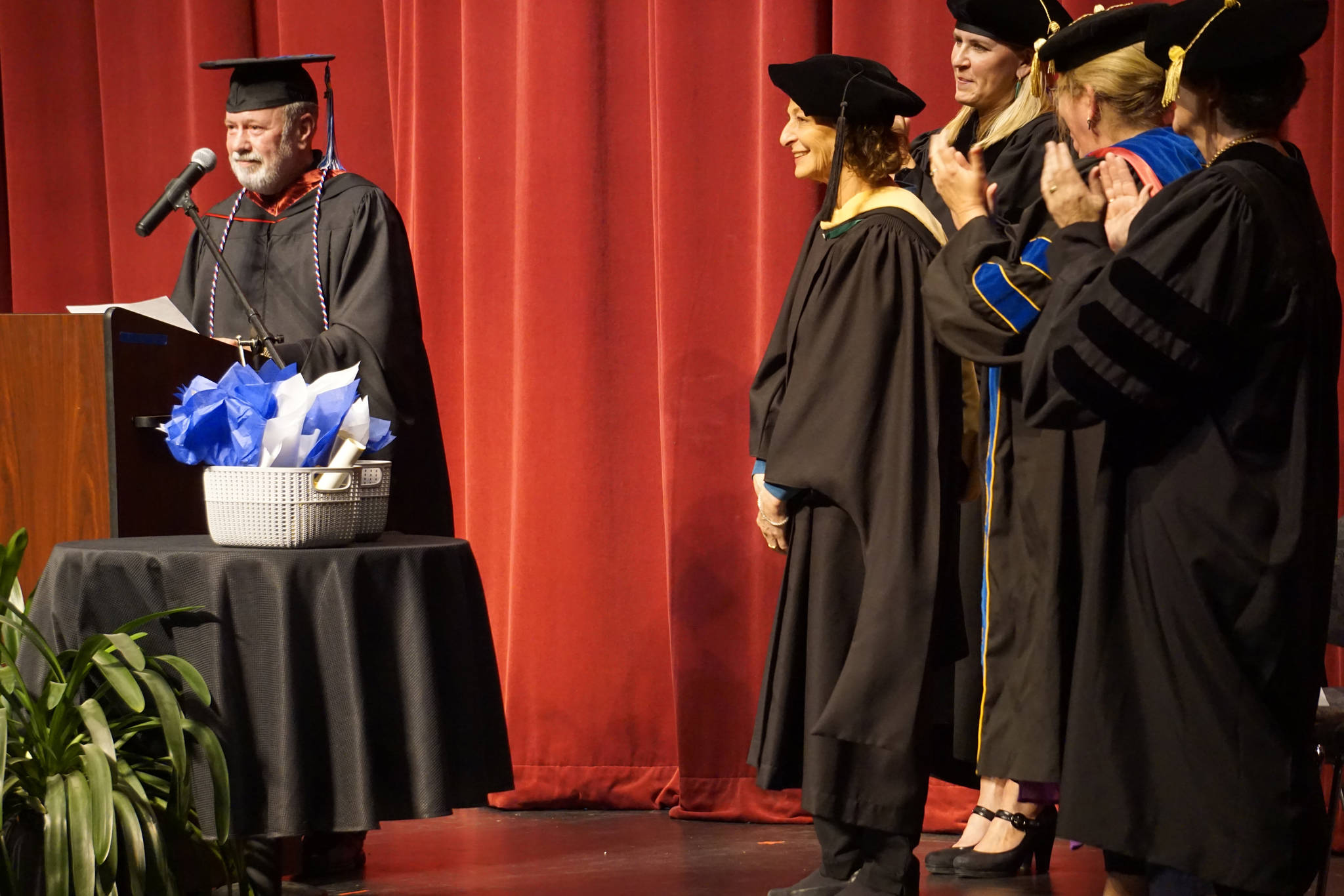 Kenai Peninsula Campus Director Gary Turner, left, reads a commendation awarding the title of Director Emerita to former Kachemak Bay Campus Director Carol Swartz, center, at the Kachemak Bay Campus commencement last Wednesday, May 8, 2019, at the Mariner Theatre in Homer, Alaska. Swartz retired last year. (Photo by Michael Armstrong/Homer News.)