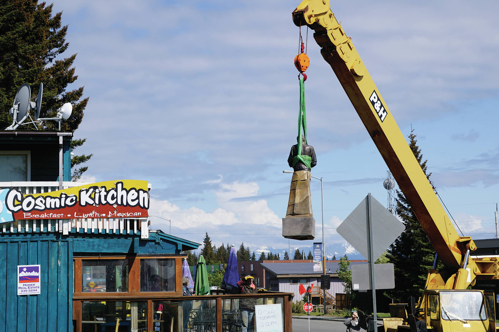 Mike Kennedy, in the crane at right, raises the Brother Asaiah statue from the deck of Cosmic Kitchen on Saturday, June 6, 2020, in Homer, Alaska. Leo Vait, left, on the deck, created the statue as a commission by John Nazarian, a friend of Asaiah Bates. Nazarian had loaned to the Pioneer Avenue restaurant the statue of the man who coined the phrase “Cosmic Hamlet by the Sea” to describe Homer. The statue was moved after Cosmic Kitchen owners Michelle Wilson and Sean Hogan sold their restaurant. Cosmic Kitchen closed on Saturday. The Asaiah statue will be stored until a new location can be found. (Photo by Michael Armstrong/Homer News)