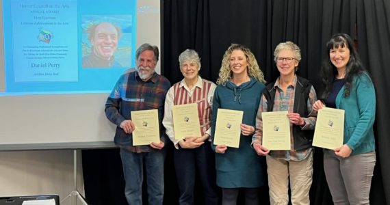 (from left to right) HCOA Community Arts awardees Daniel Perry (on screen), Michael Armstrong, Cathy Stingley, Krista Etzweiler, Sherry Stead and Brianna Allen pose with their awards at the HCOA annual meeting on Jan. 27, 2023, in Homer, Alaska.