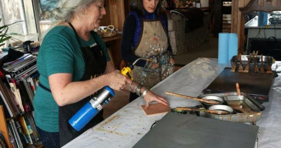 Ann-Margaret Wimmerstedt heats a plywood board for painting with encaustics on July 20, 2018 at Wimmerstedt’s home as artist Carla Klinker-Cope watches.