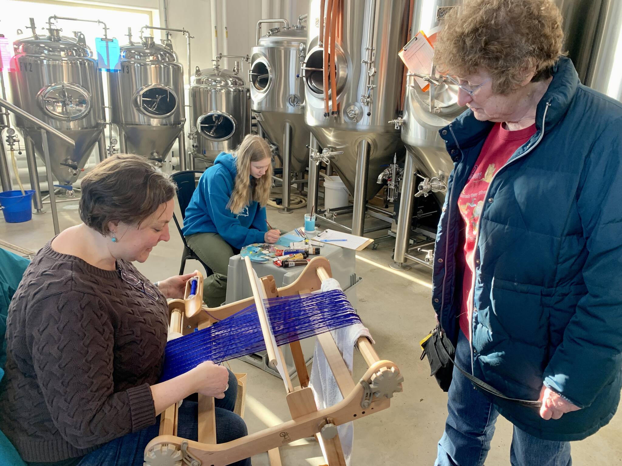 Janet Higley, right, looks on while Lisa Talbott weaves and Leah Dunn, rear center, paints during the Ptarmigan Arts scholarship fundraiser at Grace Ridge Brewing on Saturday. (Photo by Christina Whiting/Homer News)