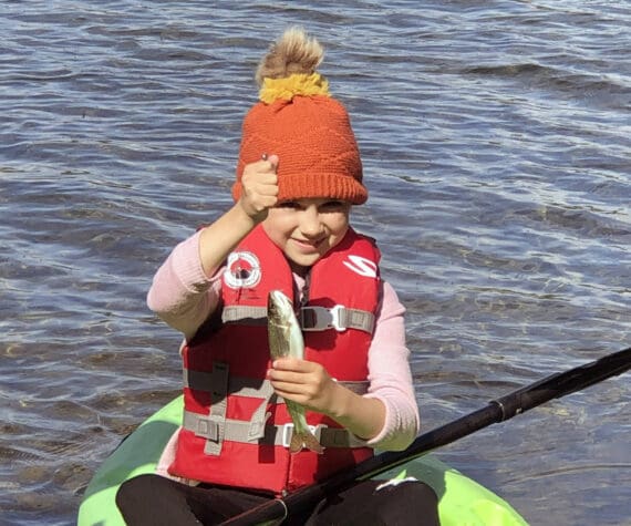 If you teach a kid to fish, she will feed you a trout breakfast every morning of the campout. This proved true for this girl, who holds up a rainbow trout while sitting on a kayak. (Photo by Leah Eskelin)