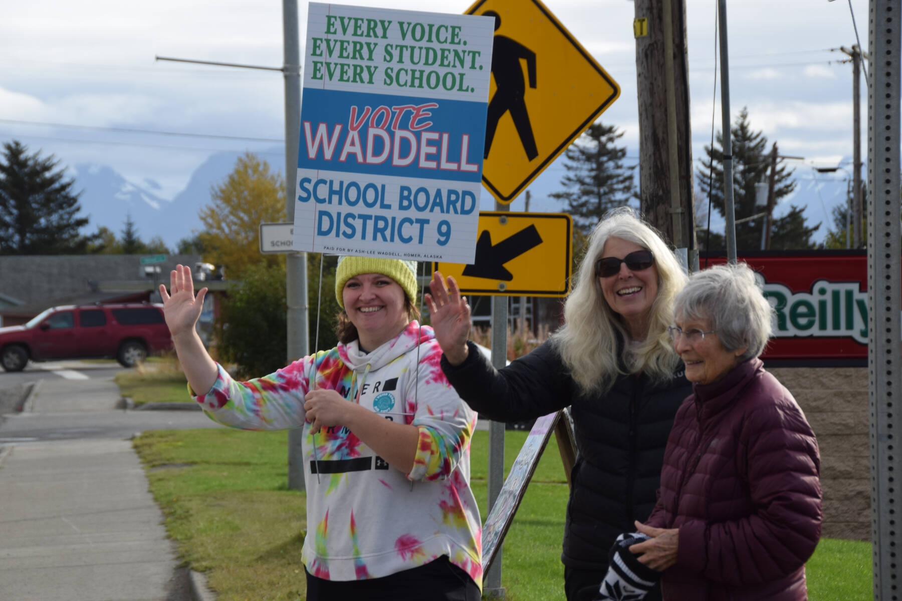 Ash-Lee Waddell (left), candidate for the District 9 seat on the Kenai Peninsula Borough School District Board of Education, holds a campaign sign and waves with supporters to passersby along the Sterling Highway in Homer, Alaska, on Election Day, Tuesday, Oct. 7, 2025. (Delcenia Cosman/Homer News)
