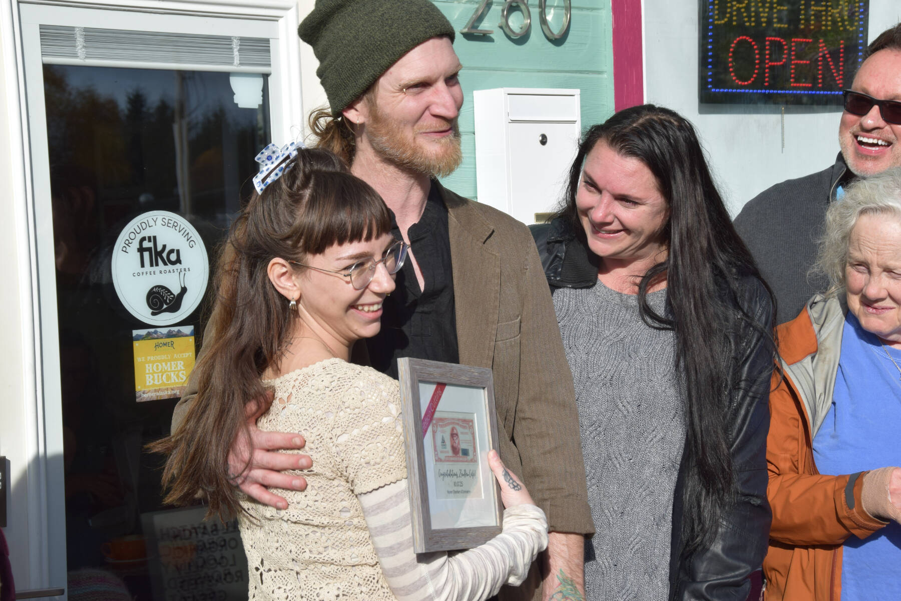 Sierra Moskios (left) and Tyler Schlieman (center), owners and operators of Fika Coffee Roasters in Homer, Alaska, hold up their first Homer Bucks bestowed on them by the Homer Chamber of Commerce during their grand opening ribbon cutting ceremony on Tuesday, Oct. 7, 2025, at ZenDen Cafe on Pioneer Avenue. (Delcenia Cosman/Homer News)