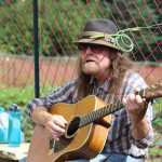 Kenute Tonga strums on his guitar and sings in the sun, adorned with a garlic scape in his hat, during Synergy Gardens' First Annual Great Garlic Scape Festival on Sunday, July 10.