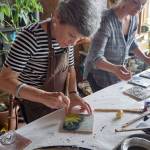 Jane Bernard of Morgan City, La., left, and Becca Bottebaum paint with encaustics in Ann-Margret Wimmerstedts Wax. Wine & Wimmerstedt class held July 30, 2017 at Wimmerstedt&rsquo;s home. (Photo by Michael Armstrong/Homer News)
