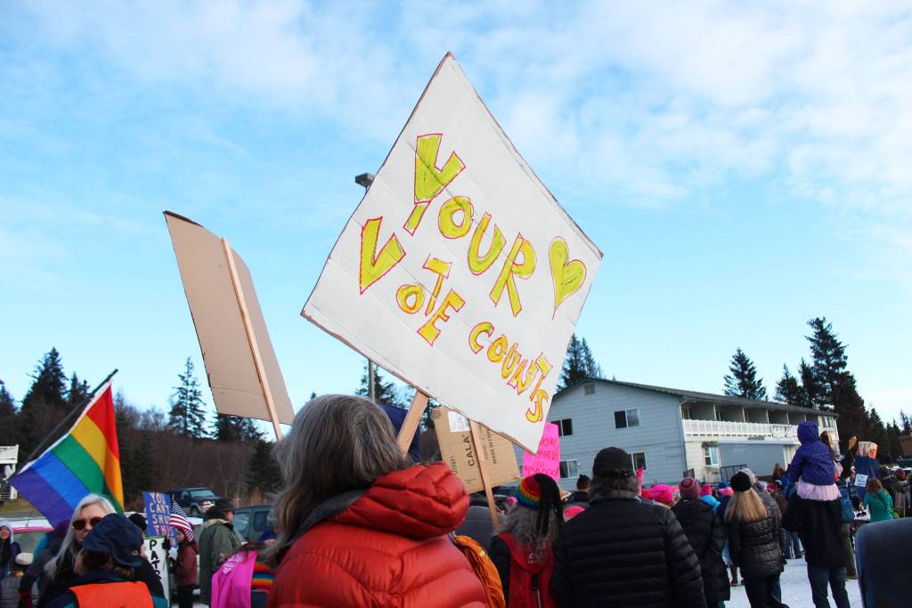 A sign reading &ldquo;Your Vote Counts&rdquo; is hoisted into the air as a crowd prepares to march in the HERC Building parking lot before the Women&rsquo;s March on Homer 2018 on Saturday, Jan. 20, 2018 in Homer, Alaska. A major focus of this year&rsquo;s march was voter registration. Registrars were set up to greet people at the end of the march in WKFL Park. (Photo by Megan Pacer/Homer News)