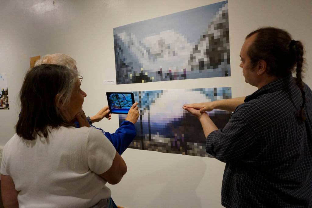 James Dolma views an image in an iPad from the Dirigibles of Denali show at the Pratt Museum at the July 6, 2018 First Friday opening in Homer, Alaska. The images appear when viewed through the HP Reveal augmented reality app. Artist Nathan Shafer, far right, explains the process to Brenda Dolma, center. (Photo by Michael Armstrong/Homer News)