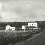 A view of Main Street looking north at the intersection of Bunnell Avenue, with, from left to right, the Homer Women's Club, the Homer Cafe and Club, and Berry's Store. The Women's Club was moved to Pioneer Avenue and was the old Homer News building before becoming Cafe Cups. The Homer Cafe and Club is now AJ's Steakhouse and Tavern, and Berry's is now the Old Inlet Trading Post building housing the Bunnell Arts Center and Maura's Cafe. The field in the foreground is where the Driftwood RV Park is now. -Photo provided; Pratt Museum; William Wakeland collection