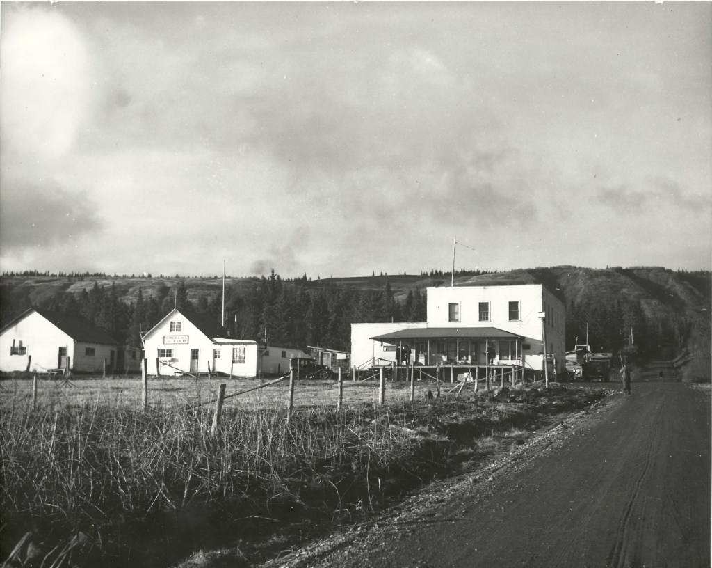 A view of Main Street looking north at the intersection of Bunnell Avenue, with, from left to right, the Homer Women's Club, the Homer Cafe and Club, and Berry's Store. The Women's Club was moved to Pioneer Avenue and was the old Homer News building before becoming Cafe Cups. The Homer Cafe and Club is now AJ's Steakhouse and Tavern, and Berry's is now the Old Inlet Trading Post building housing the Bunnell Arts Center and Maura's Cafe. The field in the foreground is where the Driftwood RV Park is now. -Photo provided; Pratt Museum; William Wakeland collection