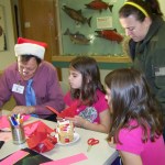 Bill Walker, exhibits assistant of the Pratt Museum, gives Paige and Morgan Haines, 8, of Homer some craft tips at the museum’s Stocking Stuffer Party on Saturday.-McKibben Jackinsky,  Homer News
