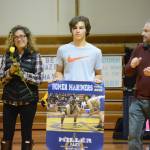 Alex Miller, center, holds a banner recognizing him at Senior Night, the Homer-Kenai dual wrestling meet held Nov. 21, 2018, in the Homer High School Alice Witte Gym, Homer, Alaska. His mother, Loreta Miller, left, and father, Brian Miller, clap for their son. (Photo by Michael Armstrong/Homer News)