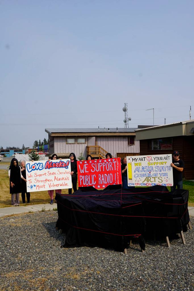 Arts supporters draped in black holds sings on July 9, 2019, by Sean Derrys public art sculpture in Homer, Alaska, as part of a statewide art intervention to protest Gov. Mike Dunleavys veto of a $2.8 million state appropriation to the Alaska State Council on the Arts. They also supported a general override of Dunleavys vetoes that will affect funding for the University of Alaska, public radio and other programs. Derrys sculpture was commissioned as a 1% for art project associated with the remodeling of Pioneer Hall at the Kachemak Bay Campus, Kenai Peninsula College, University of Alaska. The protest was not sanctioned by the college. (Photo by Michael Armstrong/Homer News)