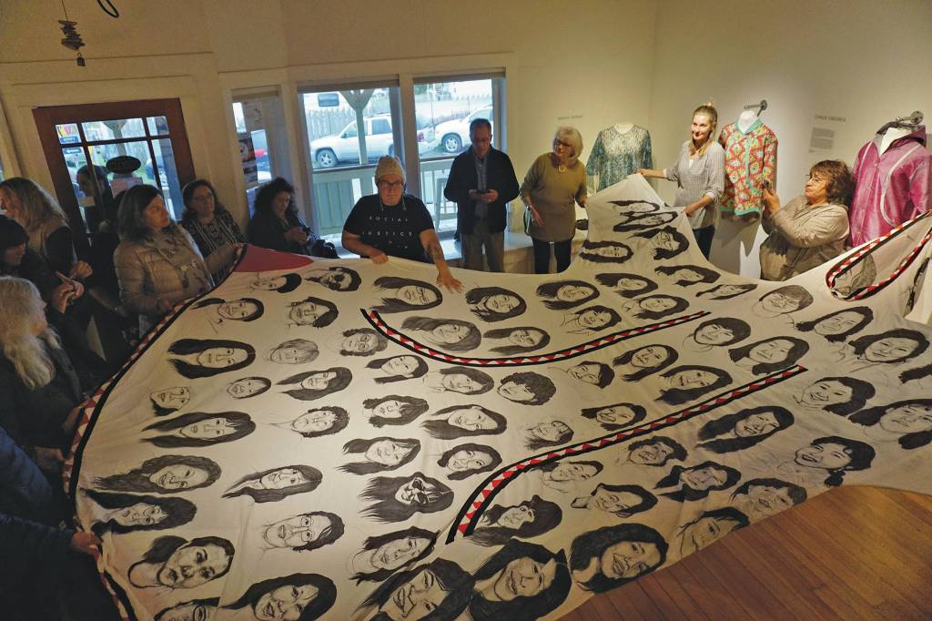 First Friday visitors hold up Amber Webbs kuspuk at the opening of the Kuspuk/Qaspeq/Atikluk show on Oct. 5, 2019, at Bunnell Street Arts Center in Homer, Alaska. The kuspuk shows faces of missing or murdered indigenous women. (Photo by Michael Armstrong/Homer News)