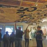 First Friday visitors hold up Amber Webbs kuspuk at the opening of the Kuspuk/Qaspeq/Atikluk show on Oct. 5, 2019, at Bunnell Street Arts Center in Homer, Alaska. The kuspuk shows faces of missing or murdered indigenous women. (Photo by Michael Armstrong/Homer News)