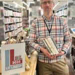 Matthew Smith, Homer Public Library staff member and the organizer for the annual Lit Lineup community reading program, poses with a selection of books next to the lineup display on Friday, Jan. 3, 2025, in Homer, Alaska. Photo by Christina Whiting