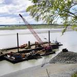 The barge, crane, and first pile of rock for the Kenai Bluff Stabilization Project is seen during a break in work at the bank of the Kenai River in Kenai, Alaska, on Tuesday, May 27, 2025. (Jake Dye/Peninsula Clarion)